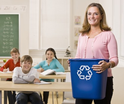 A classroom scene with a smiling young woman standing at the front, holding a blue recycling bin with white recycling symbols printed on it. Behind her, three children are seated at desks, facing forward, and appear engaged; a boy with light brown hair and a white shirt, a girl with dark hair in a light blue top, and another girl with long brown hair in a red top. The classroom features a green chalkboard on the left and large windows with white blinds letting in natural light, creating a bright atmosphere. The room has light-colored walls and wooden desks. The scene conveys a focus on environmental awareness and waste management education, aligning with services offered by Business Waste Removal Kensington for rubbish removal amidst an educational environment in London, with contextual emphasis on responsible waste practices.
