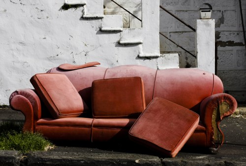 A three-seater sofa with a matching ottoman, both upholstered in medium brown leather, are placed outdoors on a paved surface. The sofa has rolled arms and a slightly worn finish, indicating frequent use, with two attached cushions on the seat and an additional cushion on the right side of the sofa. The ottoman is positioned slightly askew in front of the right armrest, with its top surface visible. In the background, there is a weathered, white-painted concrete staircase with a metal handrail, attached to a building wall that has peeling paint and dark grime. Small patches of grass grow at the base of the staircase, adjacent to the paved area. The scene appears to be part of a setup for rubbish removal or clearance of old furniture, likely in a residential or commercial setting in the Kensington area, reflecting the company's service of removing bulky waste materials like furniture for disposal or recycling.