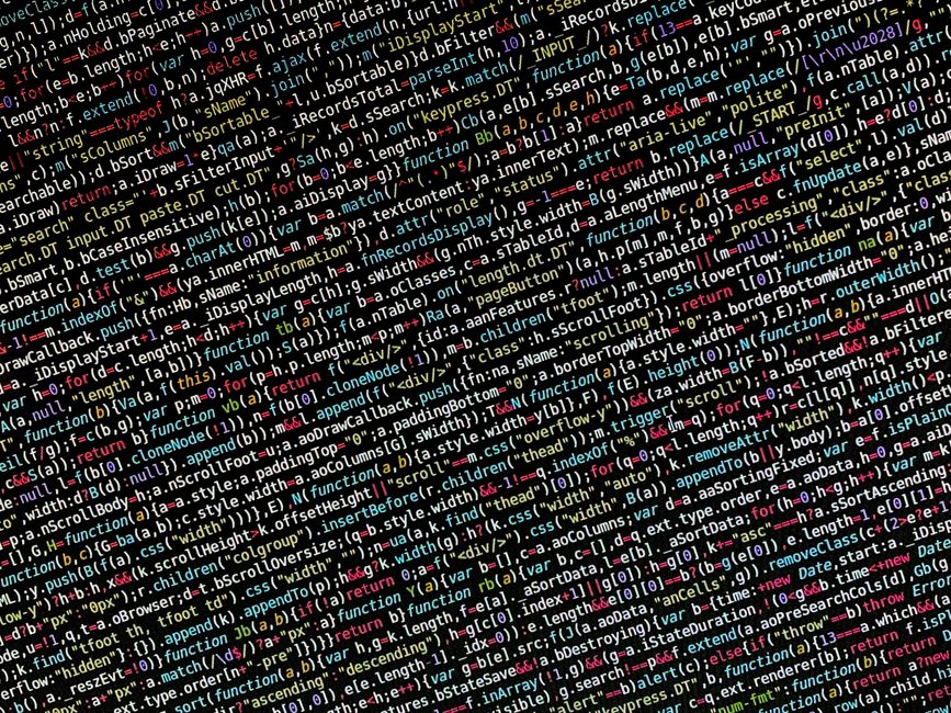 In the photograph, a close-up view of a computer screen displaying lines of colourful programming code is visible, with a mixture of text, symbols, and syntax highlighting in shades of red, green, yel