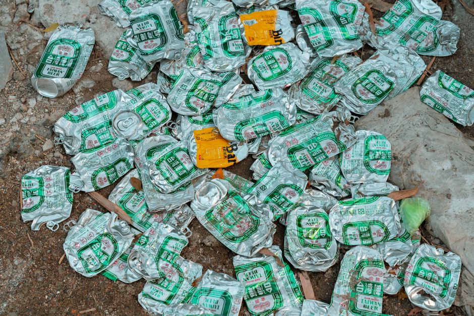 A narrow urban alleyway filled with discarded waste and rubbish. In the foreground, there is a large, black garbage bag wrapped in clear plastic, placed on a wheelie bin or trolley, with visible creas